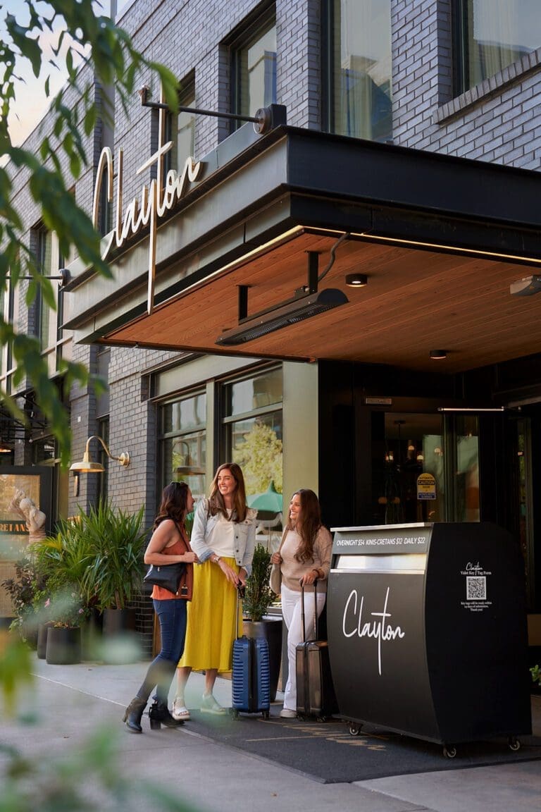 Three women outside The Clayton hotel with luggage. Hotel valet sign visible.