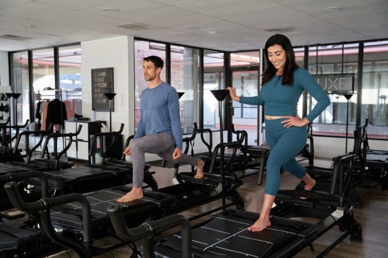 Man and woman doing Pilates on reformers in a bright studio.